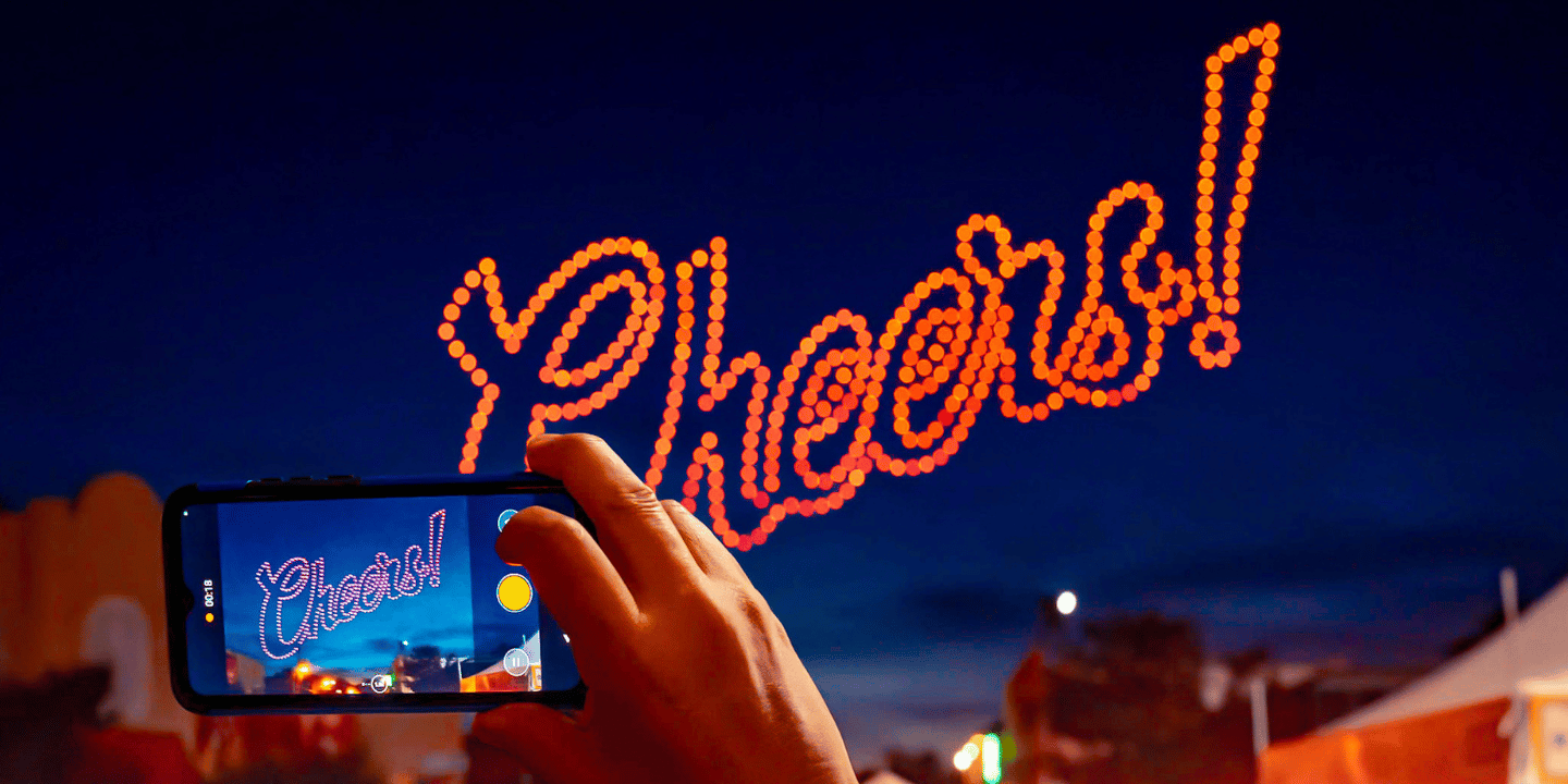 Cheers! displayed in night sky by Sky Elements Drone Shows with hand holding camera taking a picture