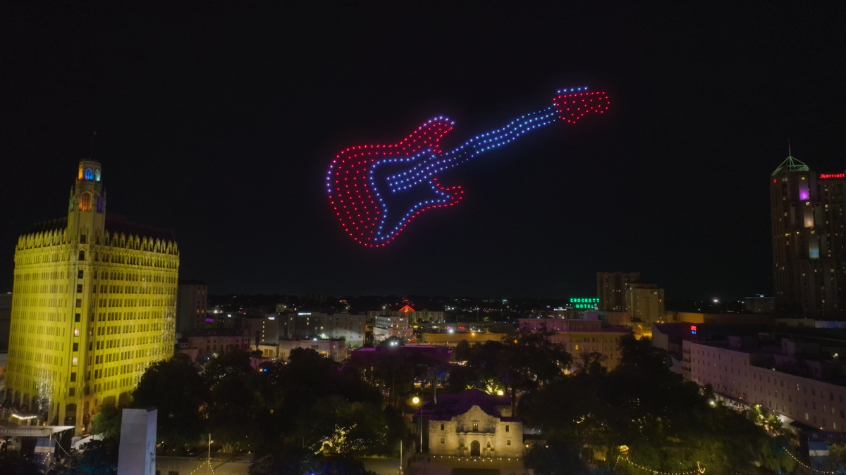 A drone light show forms a glowing red and blue electric guitar above a city skyline at night, highlighting a music-themed celebration.