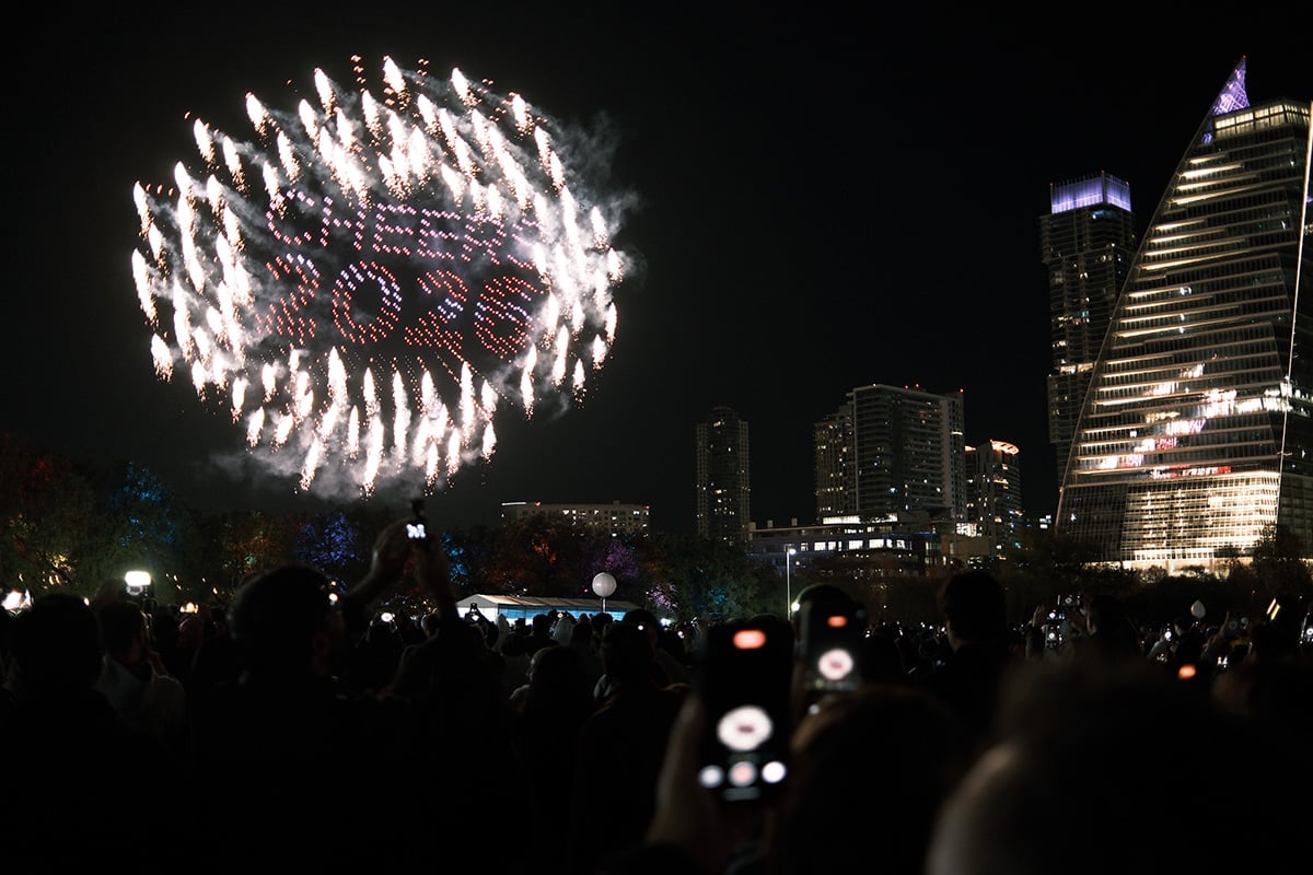 a group of people are taking a photo of drone show in downtown austin. pyro drones outline the drone show that say 