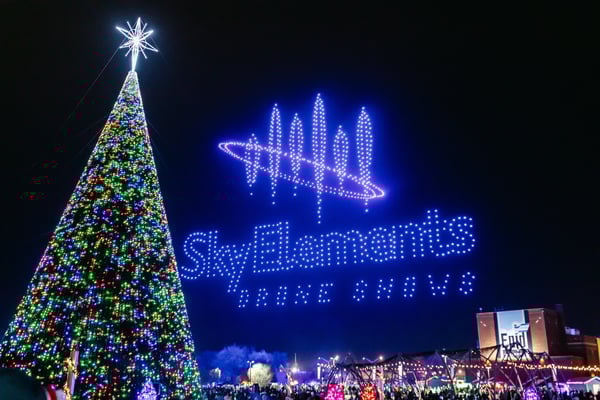 A drone light show next to a Christmas tree showing the sky elements logo