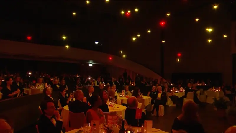 Gala audience at candlelit tables beneath warm drone lights indoors
