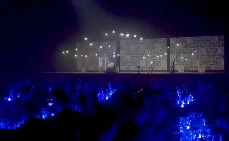Speaker on a lit stage above a blue-lit gala audience with drones overhead