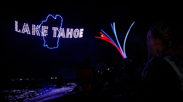 A drone light show over Lake Tahoe displays the lake's outline and name in glowing blue lights as spectators watch from the shoreline.