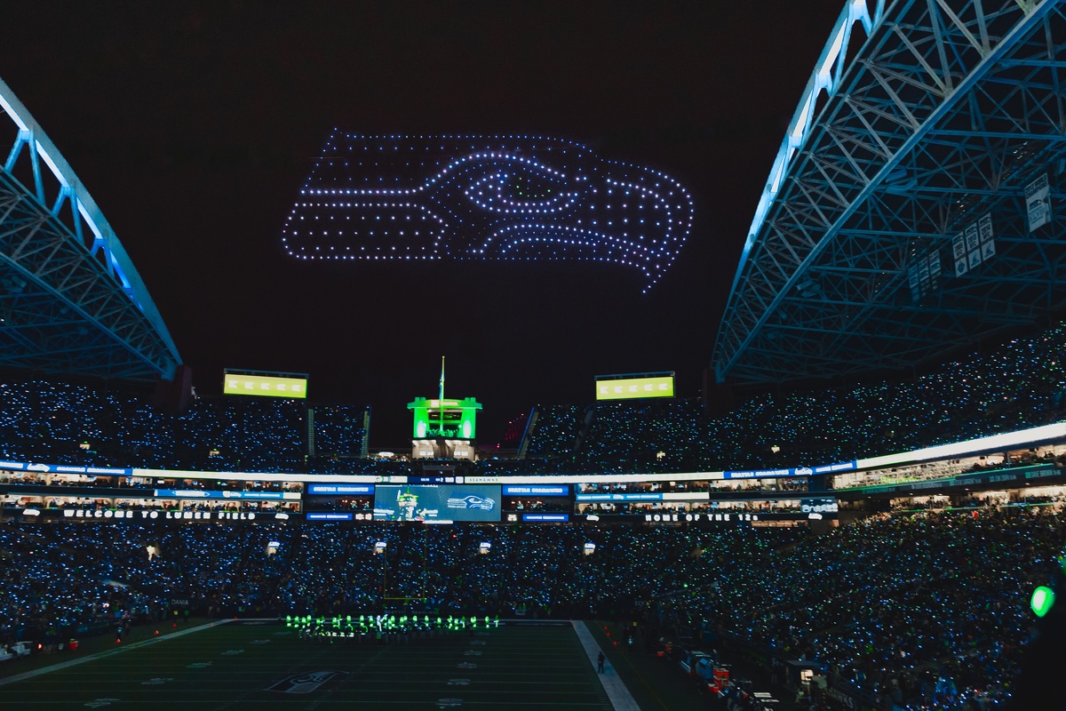A stunning drone light show forms the Seattle Seahawks logo over a packed stadium during a nighttime NFL game, illuminating the sky above the cheering crowd.